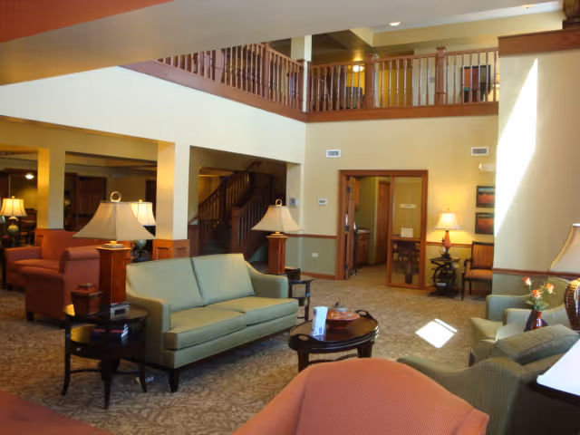 A spacious and well-lit living room area in a senior living facility featuring multiple upholstered sofas and armchairs arranged around wooden coffee tables. The room has warm beige walls, carpeted floors, several table lamps providing soft lighting, and a wooden railing on the upper level overlooking the space.