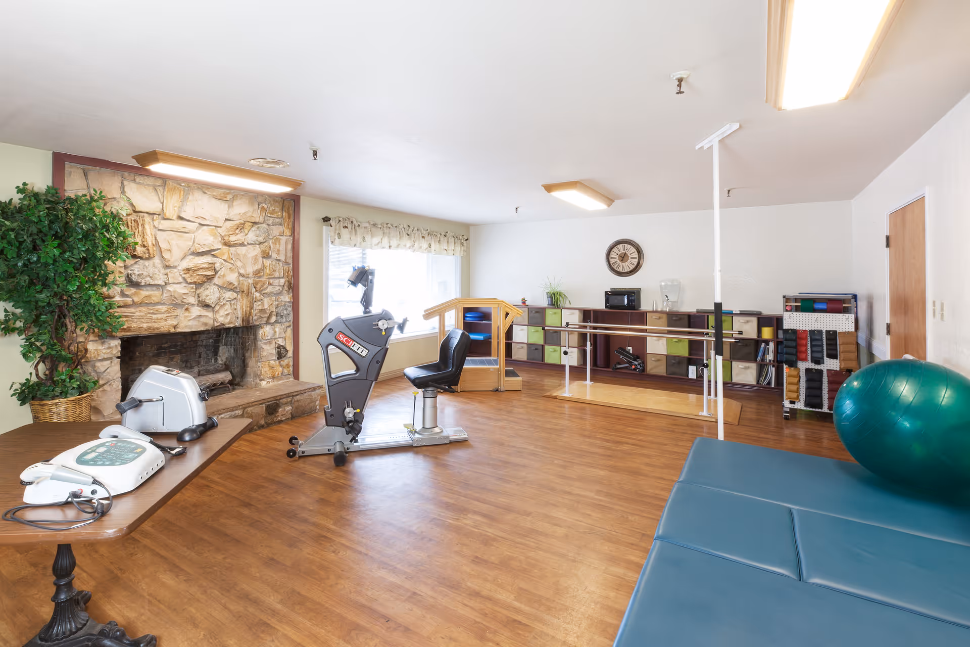 A spacious therapy or exercise room with wooden flooring, a stone fireplace on the left, exercise equipment including a stationary bike and a large green exercise ball, parallel bars for walking support, a table with medical devices, and a shelving unit with colorful storage bins against the back wall. The room is well-lit with ceiling lights and natural light from a window with a floral valance.