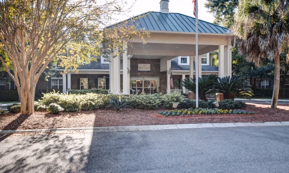 Front exterior view of a senior living facility with a covered entrance supported by white columns, surrounded by landscaped bushes, palm trees, and a flagpole with an American flag.
