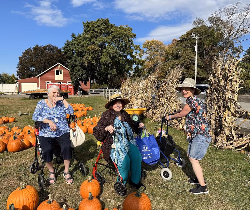 Three elderly women outdoors at a pumpkin patch on a sunny day. Two women are seated on walkers among numerous pumpkins on the grass, while the third woman stands with a walker. They are all smiling and wearing casual clothing and hats. Behind them are dried corn stalks and a red barn with a blue sky above.