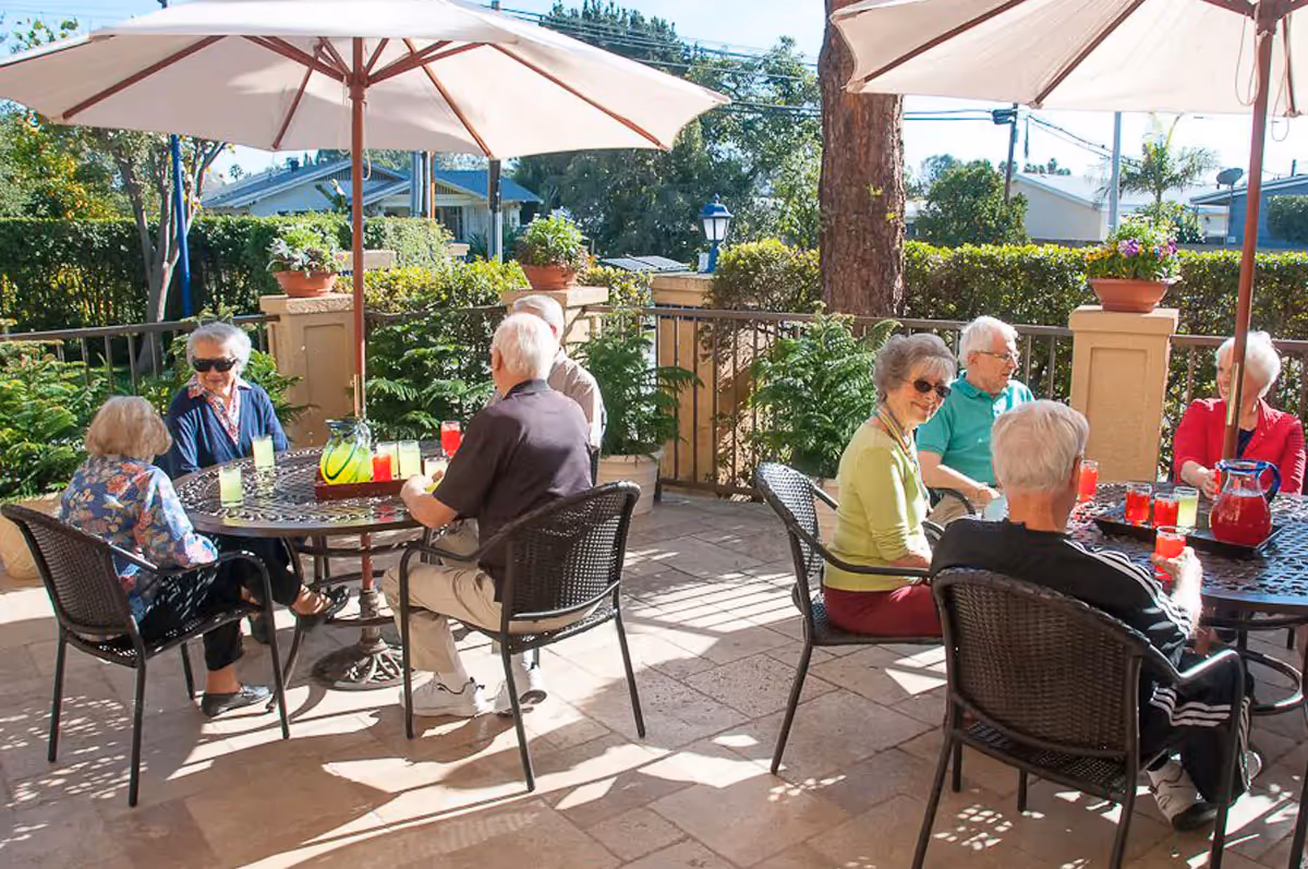 A group of elderly people sitting around two outdoor tables under large white umbrellas, enjoying drinks on a sunny patio surrounded by greenery and potted plants.