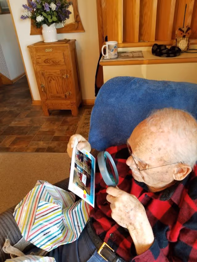 An elderly man wearing glasses and a red and black checkered shirt is sitting in a blue armchair, using a magnifying glass to closely examine a photograph. A colorful striped bag is on his lap. In the background, there is a wooden cabinet with a white vase of purple flowers on top and a wooden shelf with a mug and decorative items.