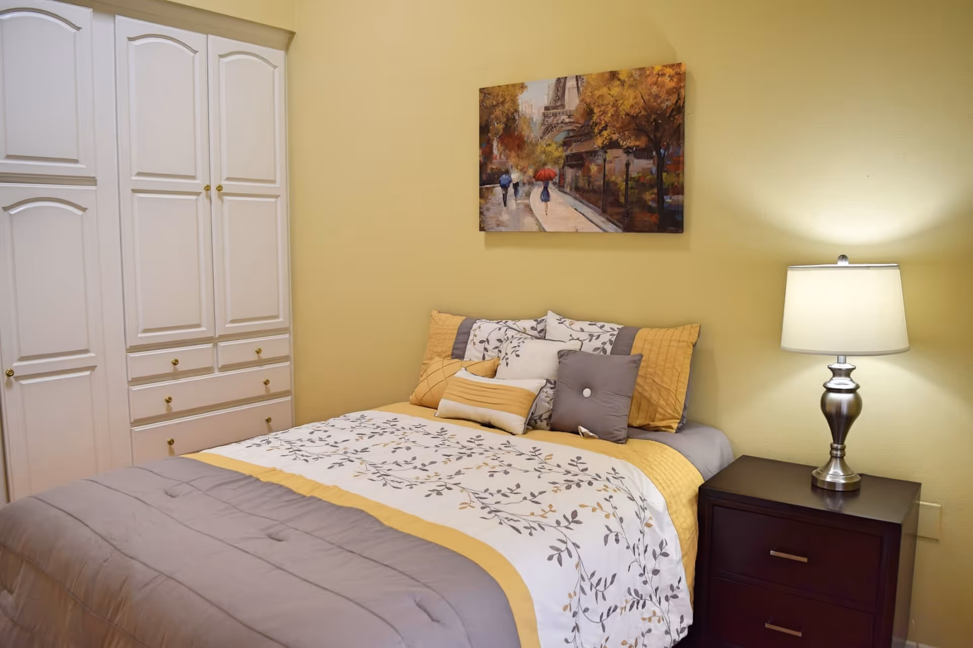 A cozy bedroom with a neatly made bed featuring a floral and yellow patterned comforter and multiple pillows. To the right of the bed is a dark wooden nightstand with a silver lamp emitting warm light. On the left side, there is a white wardrobe with multiple drawers and cabinets. A painting depicting a street scene with people holding umbrellas and autumn trees hangs on the yellow wall above the bed.