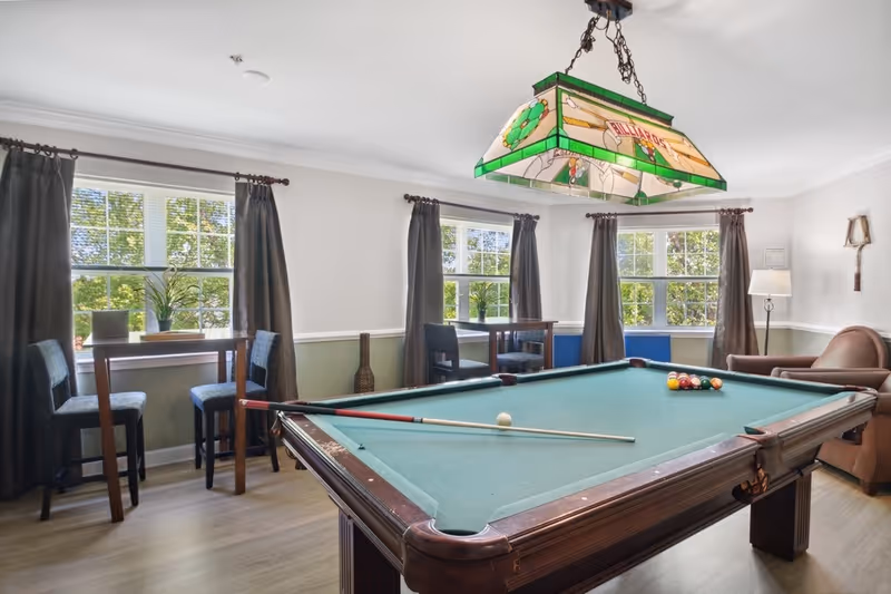 A bright room with a pool table in the center, a pool cue resting on the table, and a rack of billiard balls arranged. The room has three windows with dark curtains, two small tables with chairs near the windows, a brown armchair, a floor lamp, and a decorative wall-mounted item. A stained glass billiards-themed light fixture hangs above the pool table.