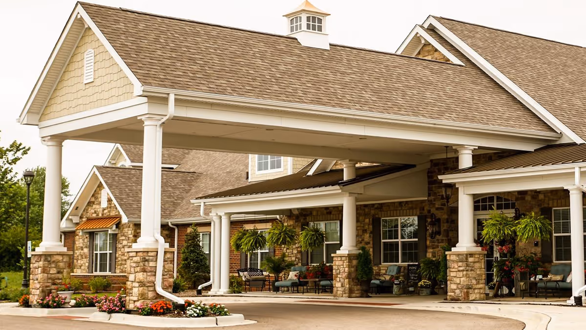 Exterior view of a senior living facility with a large covered entrance supported by white columns and stone bases. The building features a combination of brick and stone walls, multiple windows, and outdoor seating areas with plants and flowers around the entrance.