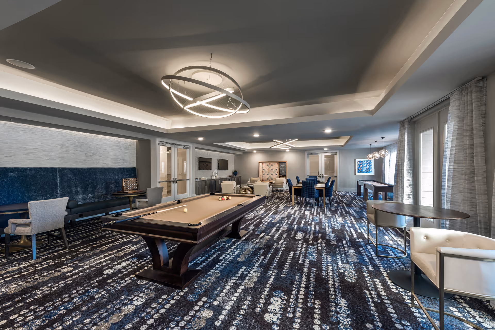 A spacious and modern game and lounge room featuring a pool table in the center, multiple seating areas with chairs and tables, a shuffleboard table near the windows, and contemporary lighting fixtures on the ceiling. The room has patterned carpet flooring and large windows with sheer curtains allowing natural light to enter.