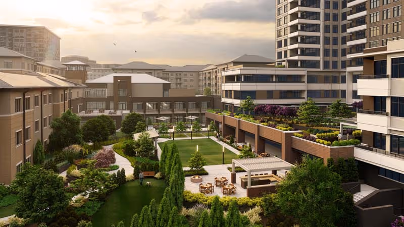 A landscaped outdoor courtyard area in a senior living facility with walking paths, green lawns, trees, shrubs, seating areas including tables and chairs, and a covered pergola. Surrounding the courtyard are multi-story residential buildings with balconies and large windows under a partly cloudy sky at sunset.