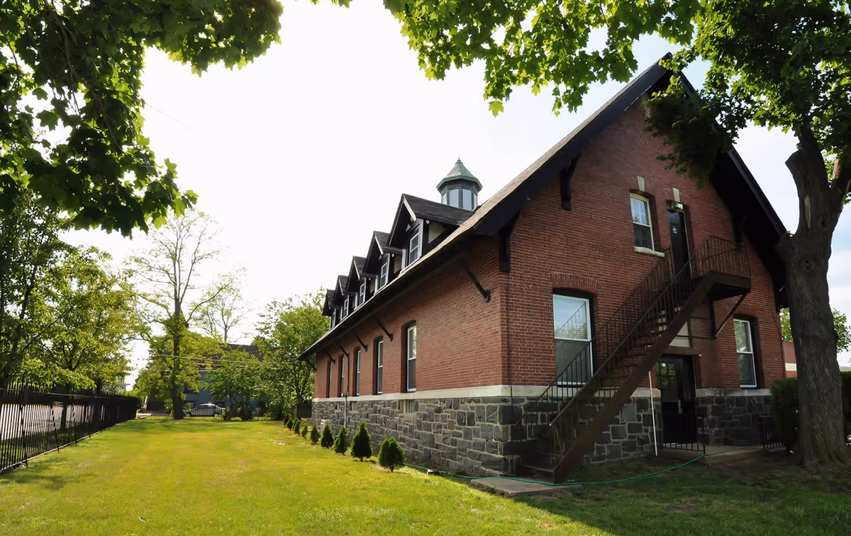Brick two-story building with an exterior metal staircase, stone foundation, green lawn, and surrounding trees.