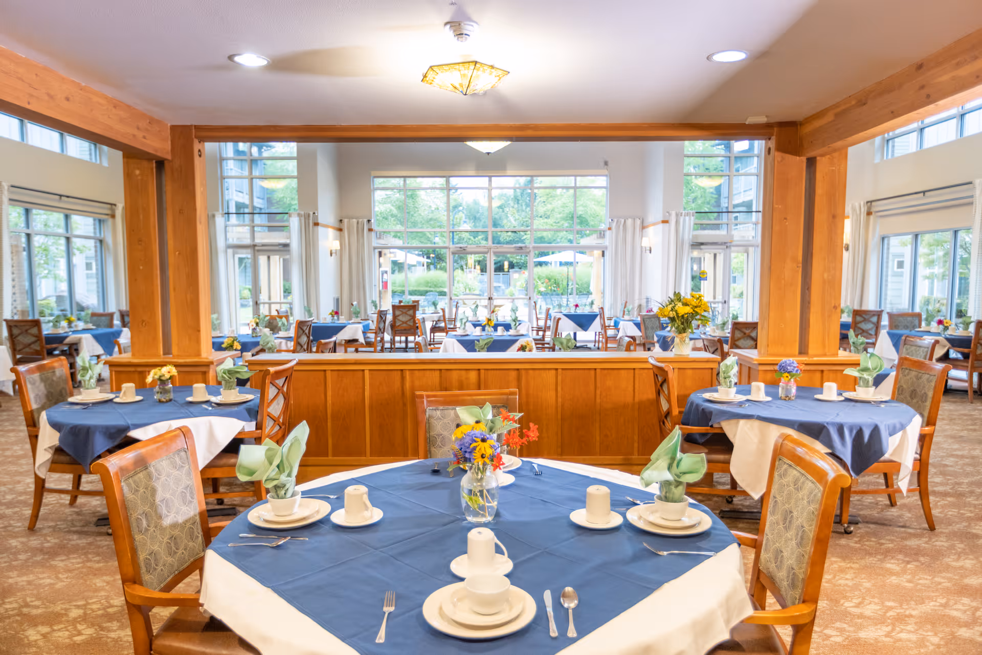 A bright and spacious dining room with multiple round tables covered in blue and white tablecloths. Each table is set with white plates, cups, silverware, and green folded napkins. There are floral centerpieces on each table. Large windows in the background let in natural light and offer a view of greenery outside. The room features wooden beams and columns, and upholstered chairs surround the tables.