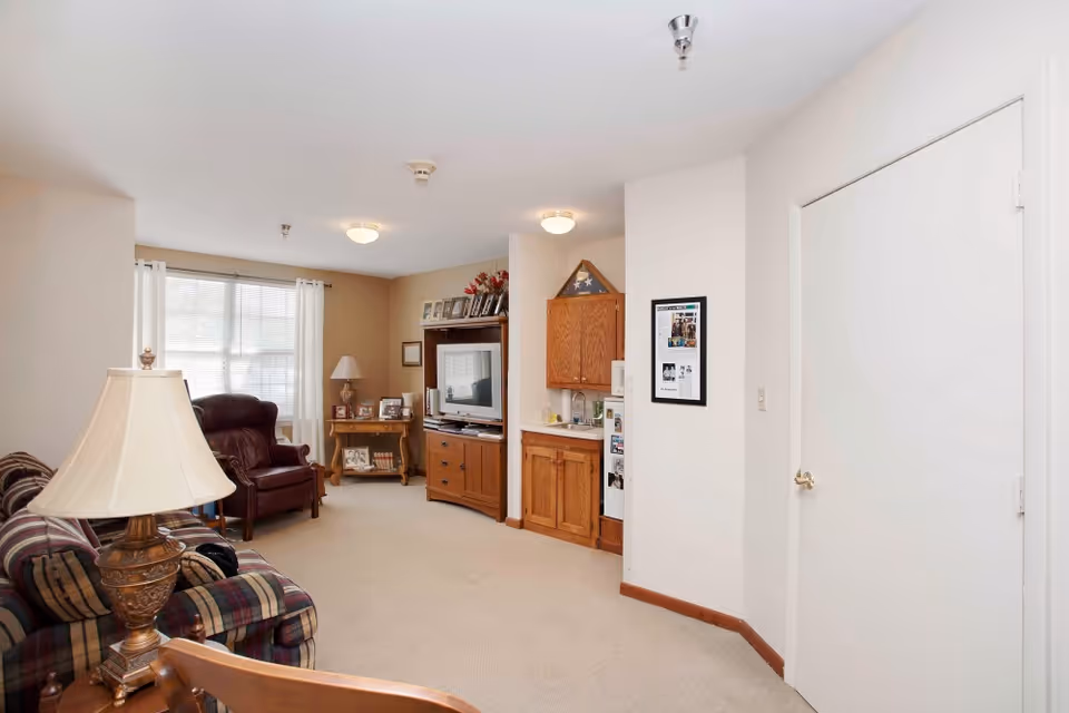 Comfortable living room with a plaid sofa and lamp in the foreground, a leather armchair by a window, and a TV cabinet and small kitchenette area along the far wall.