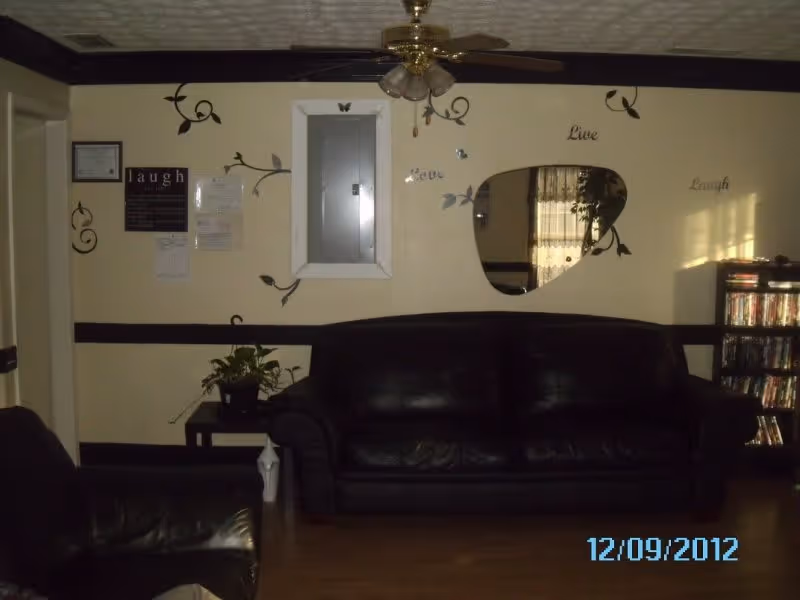 A living room with a black leather sofa and a matching armchair. The walls are decorated with black vine-like decals and words such as 'Live', 'Love', and 'Laugh'. There is a uniquely shaped mirror on the wall reflecting a window with curtains. A ceiling fan with lights hangs from the ceiling. A small table with a potted plant is beside the sofa, and a bookshelf filled with DVDs is visible on the right side. The floor is wooden, and the room has a cozy, lived-in feel.