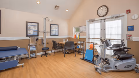 A physical therapy or rehabilitation room with exercise equipment including a recumbent bike, parallel bars, and a treatment table. There is a round table with chairs in the center, medical posters on the walls, and a clock above double doors with sheer curtains letting in natural light.