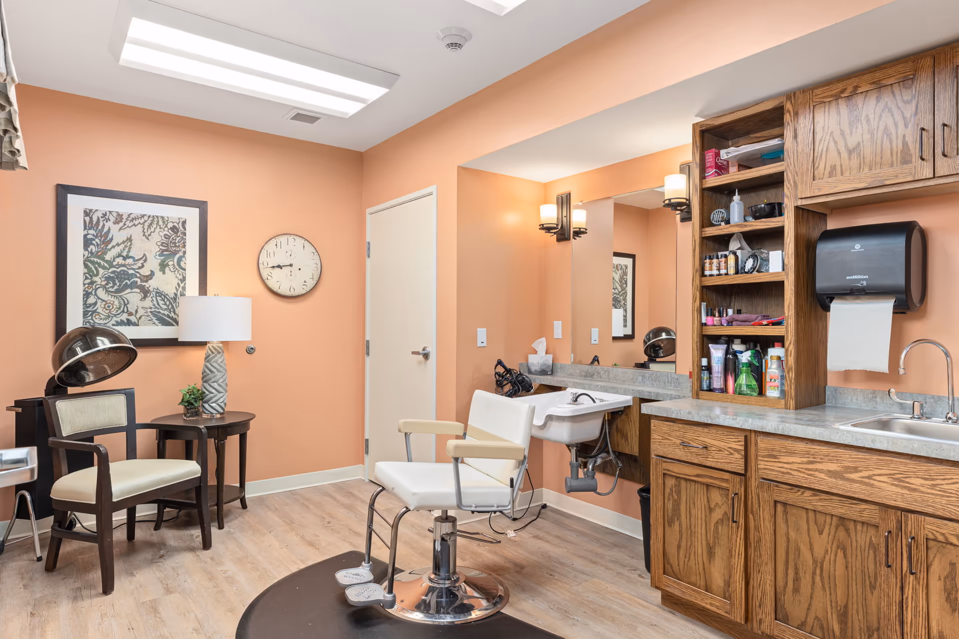Interior view of a hair salon area in a senior living facility with peach-colored walls, a white salon chair in front of a sink, wooden cabinets with various hair care products, a wall-mounted paper towel dispenser, a small round table with a lamp and plant, a vintage hair dryer chair, a framed floral artwork, and a wall clock.