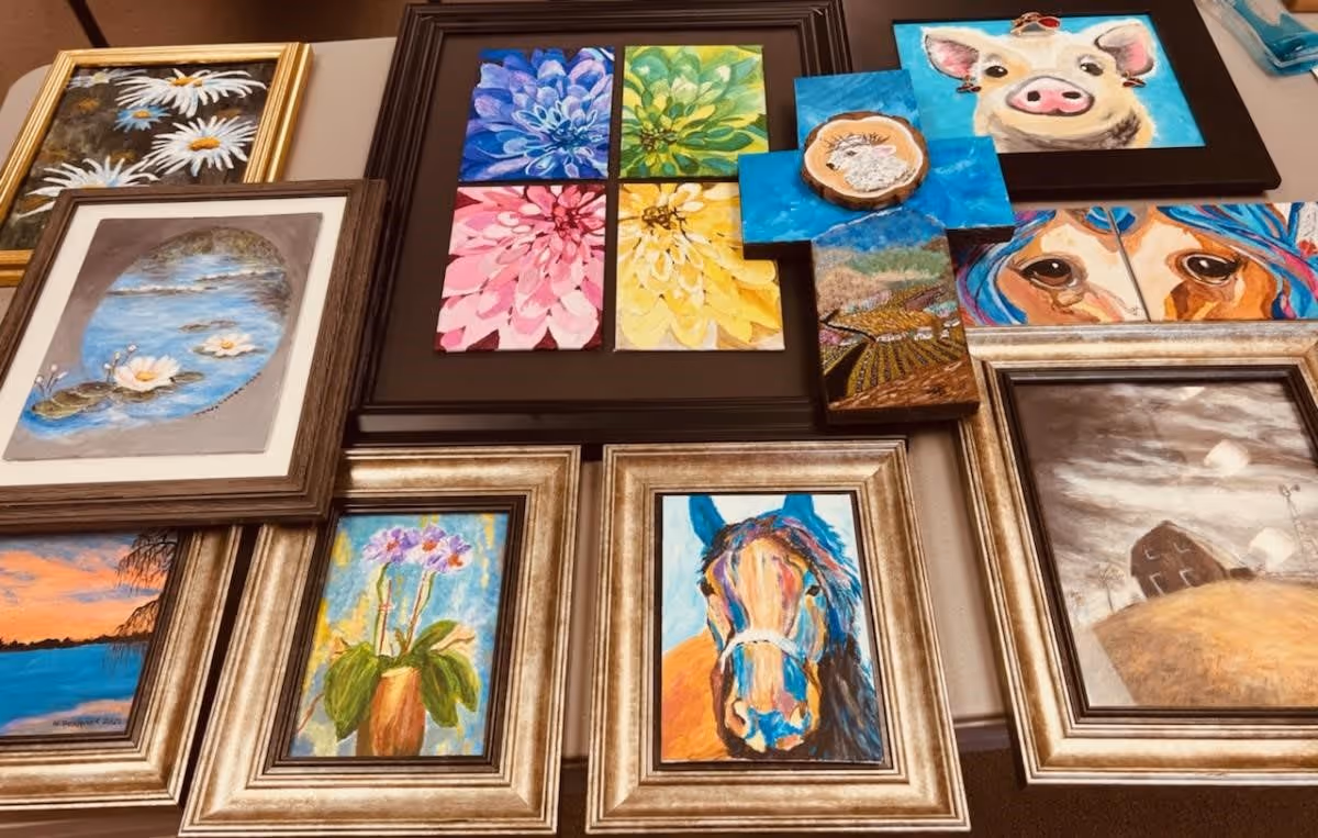 A collection of framed paintings displayed on a wall, featuring various subjects including flowers, a pig, a horse, a dog, a landscape with a barn, and a cross with a small painting of a sheep on wood.