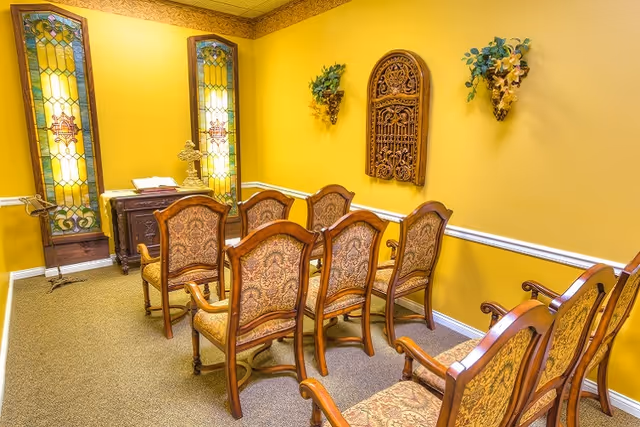 A small chapel or prayer room with yellow walls, stained glass windows, and ornate wooden chairs arranged in rows facing a small altar with a cross and an open book.