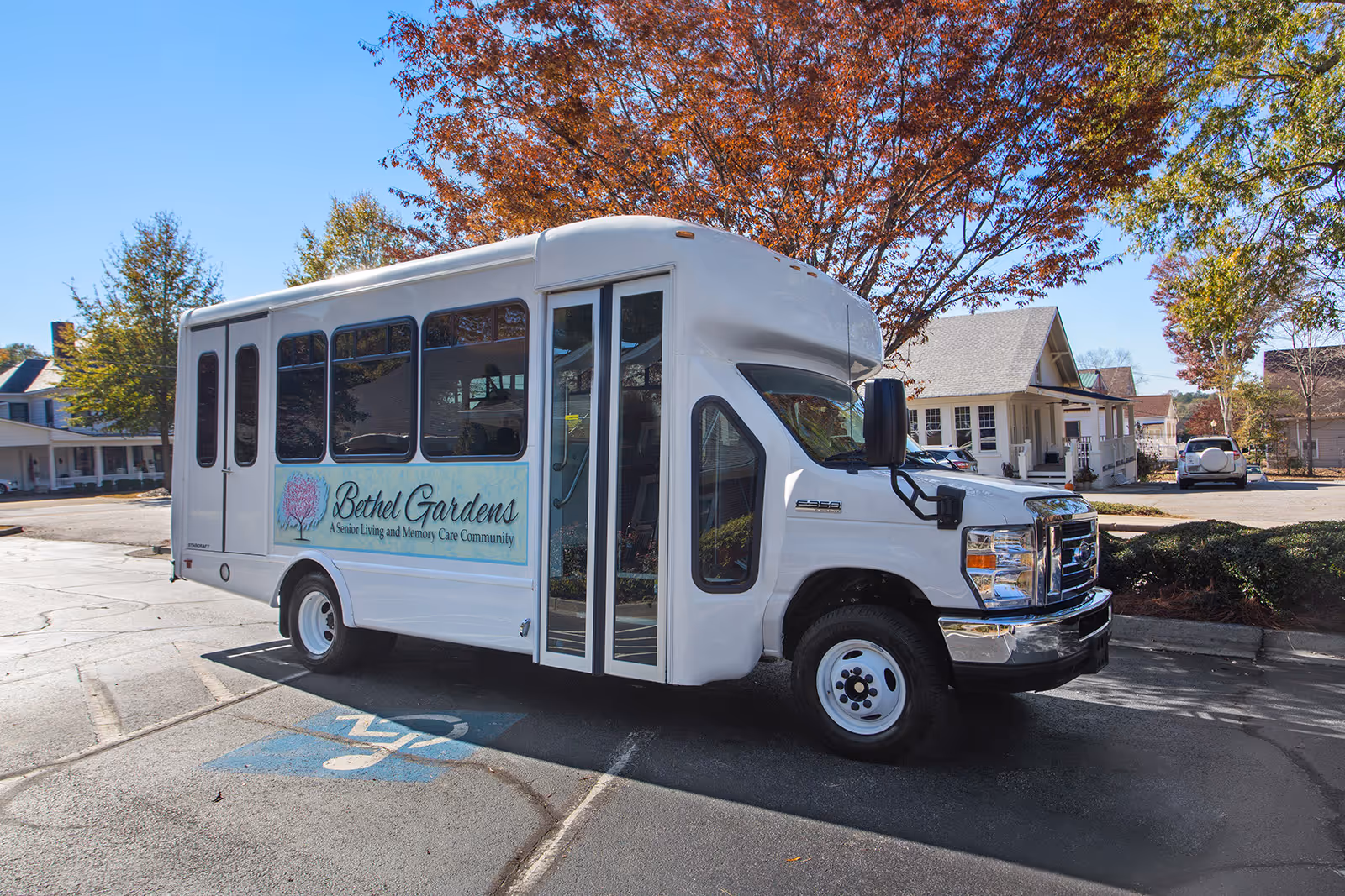 A white shuttle bus parked in a parking lot with a handicap parking space visible in front. The bus has a sign on the side that reads 'Bethel Gardens A Senior Living and Memory Care Community'. Trees with autumn-colored leaves and residential buildings are visible in the background under a clear blue sky.
