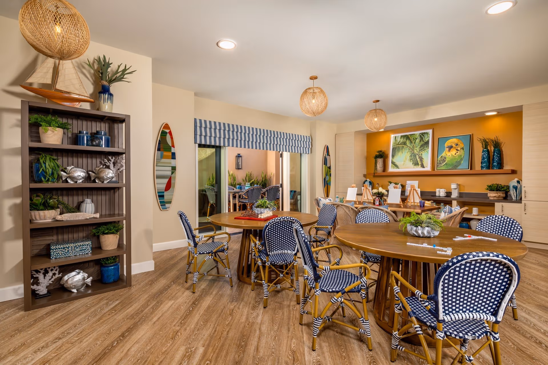 A bright and inviting activity room with round wooden tables surrounded by blue and white woven chairs. The room features wooden flooring, a shelf with decorative plants and fish sculptures, two surfboard-shaped wall decorations, and a sliding glass door leading to an outdoor patio. The back wall has a mustard yellow accent with framed pictures of tropical scenes and plants, and several small easels set up for painting or crafts.