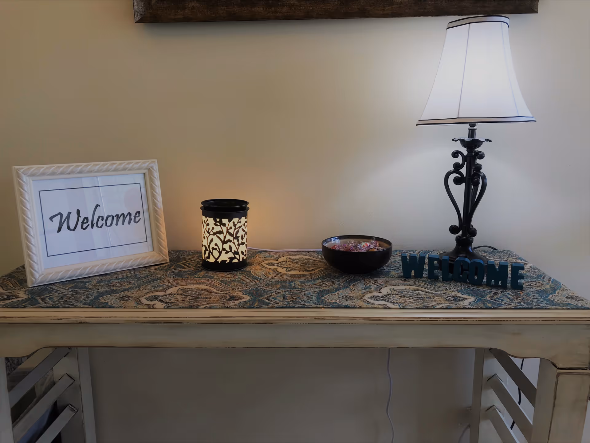 A decorative table with a patterned runner, a framed sign that says 'Welcome', a lit decorative candle holder, a bowl with wrapped candies, a black metal 'WELCOME' sign, and a table lamp with a white shade against a plain wall.
