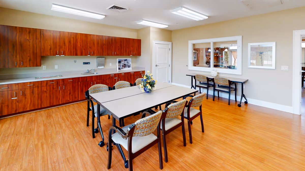 A bright room with wooden flooring and beige walls featuring a long table with six chairs around it. There are wooden cabinets along one wall with a countertop, a sink, and a paper towel dispenser. On the opposite wall, there is a window looking into another room and a small desk with two chairs. The room is well-lit with ceiling lights.