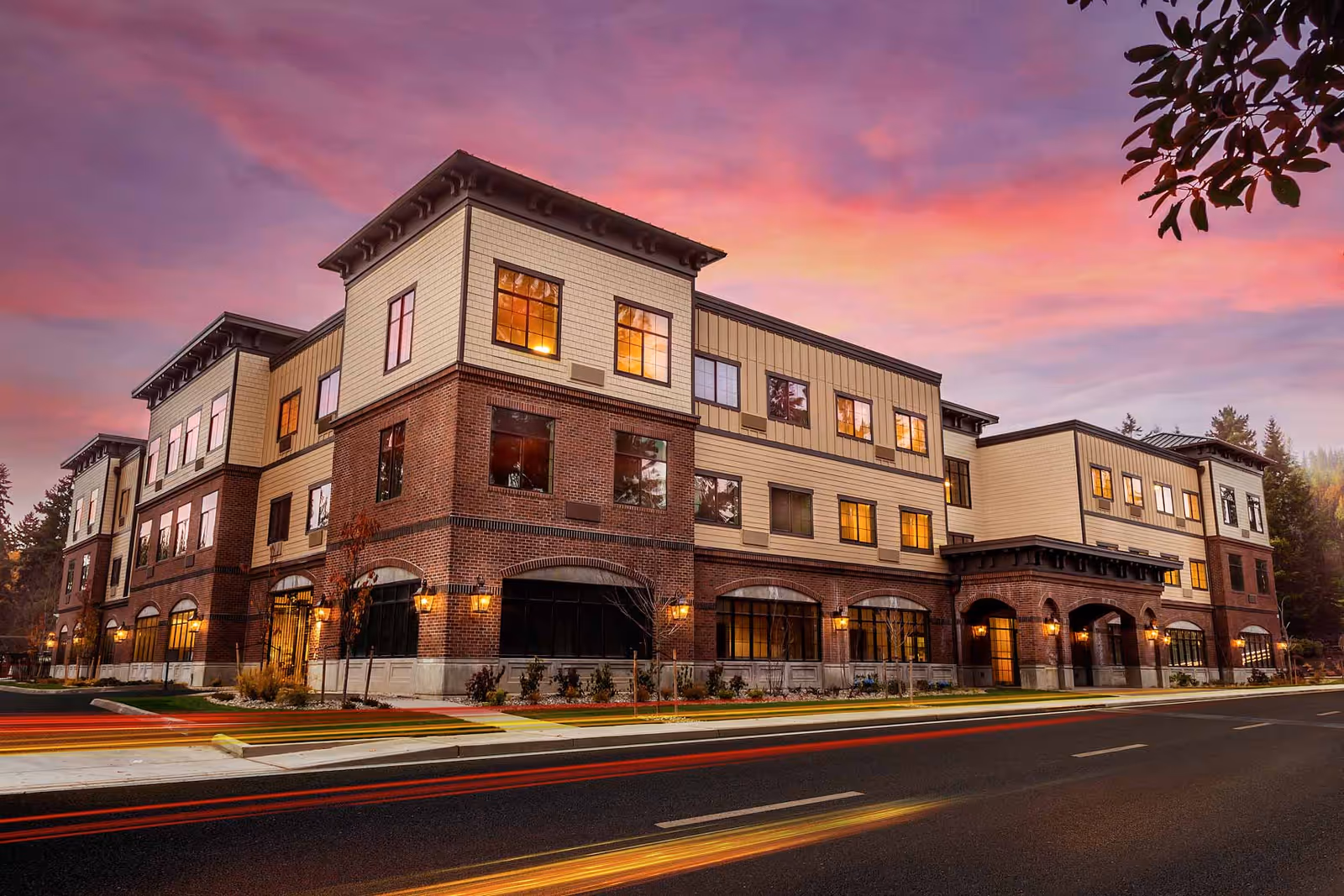 Exterior view of a three-story senior living facility building with brick and beige siding under a colorful sunset sky. The building has multiple windows with warm lights glowing inside and is situated next to a road with light trails from passing vehicles.