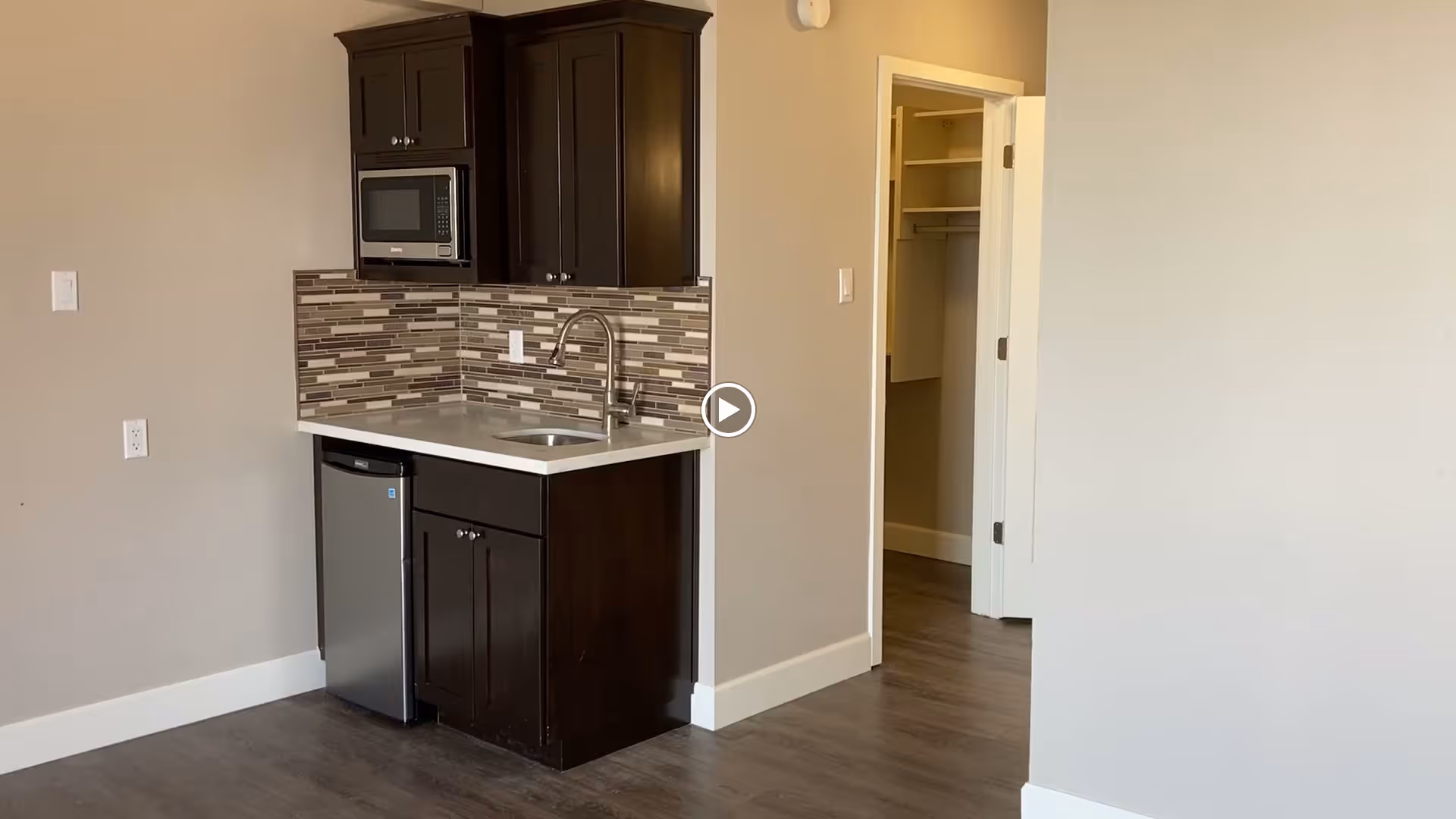Compact kitchenette with dark wood cabinets, microwave, sink, mini refrigerator and tile backsplash beside an open doorway to a closet.