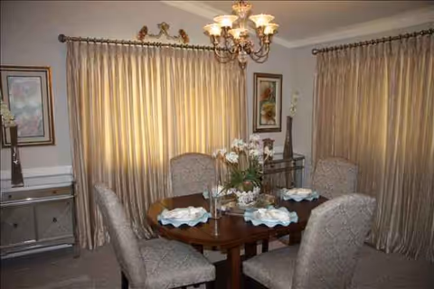A formal dining room with a round wooden table, six upholstered chairs, chandelier, and draped windows.