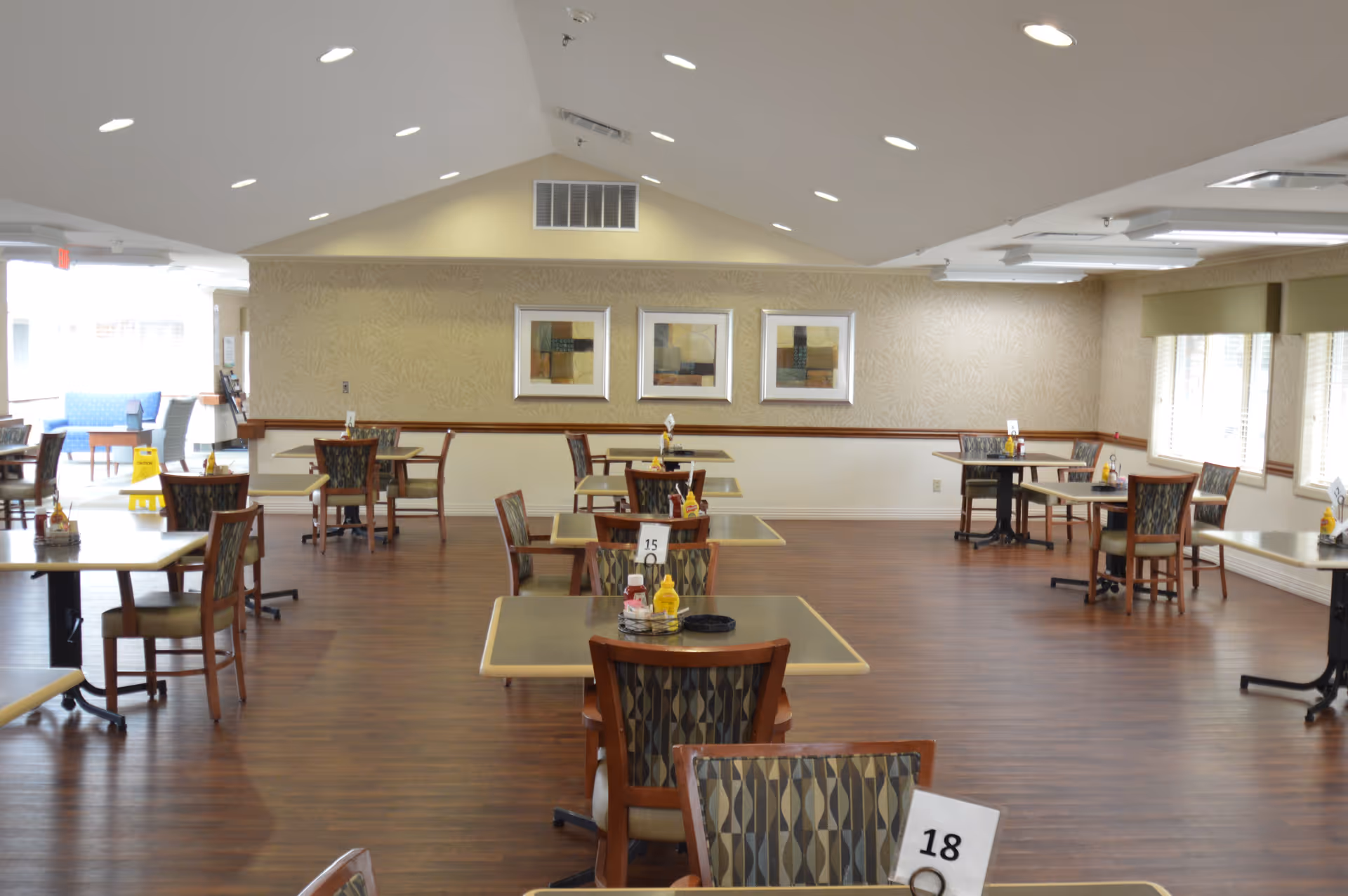 A spacious dining room with multiple tables and chairs arranged neatly. Each table has condiments and a table number. The room has wooden flooring, beige walls with three framed abstract paintings, and large windows with blinds allowing natural light to enter.