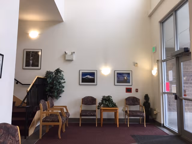 Entrance lobby with seating: several chairs around a small table, potted plants, framed pictures on a tall white wall, and glass entry doors.