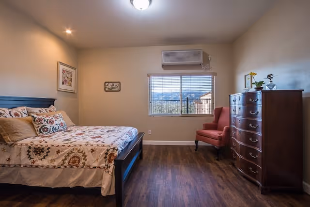 A bedroom with a wooden bed frame and floral patterned bedding, a red upholstered armchair near a window with blinds, a wooden dresser with decorative items on top, and hardwood flooring. The walls are beige and there is an air conditioning unit above the window.