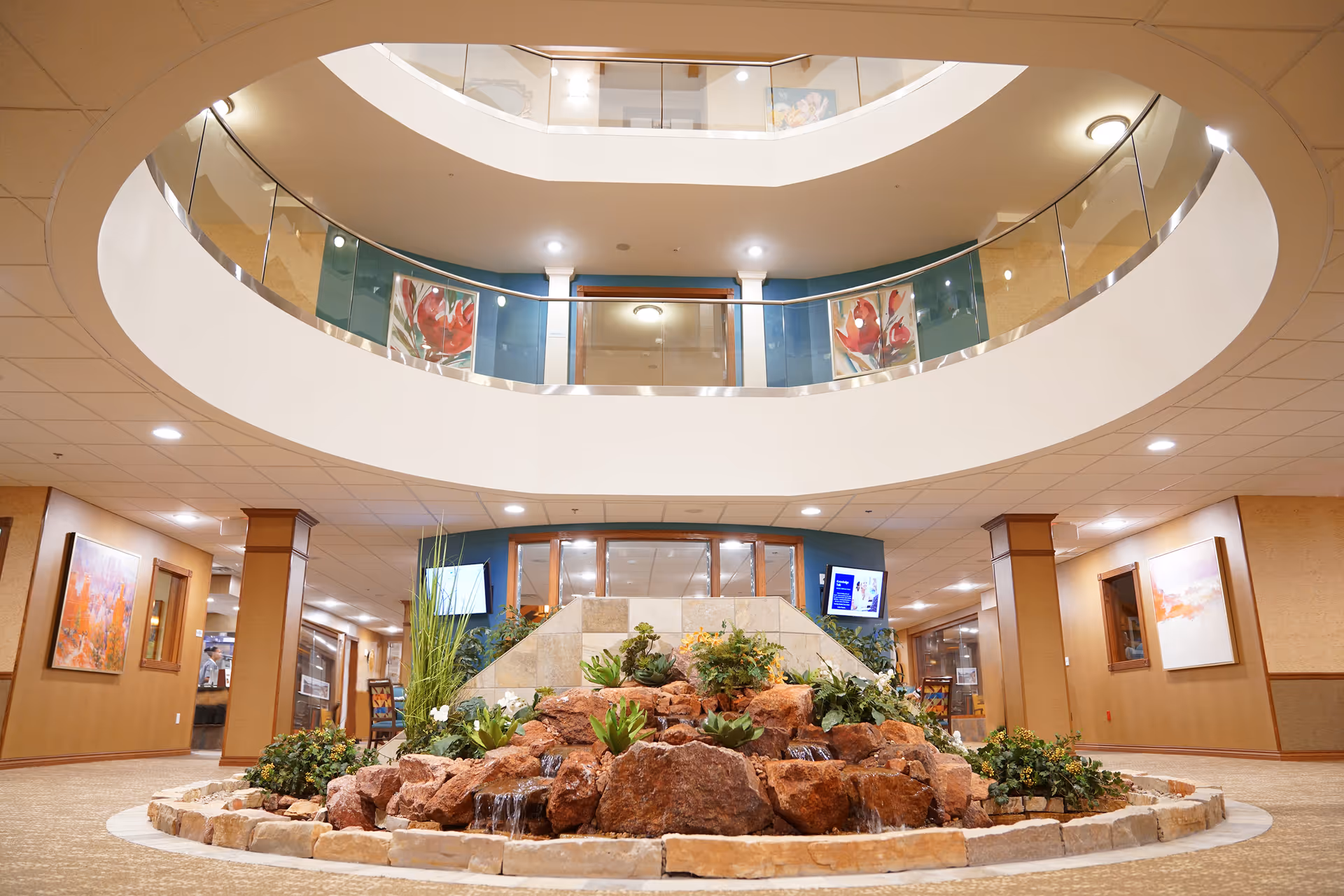 Wide interior atrium of a retirement facility featuring a central rock and plant water feature beneath a two-story circular balcony with glass railings.