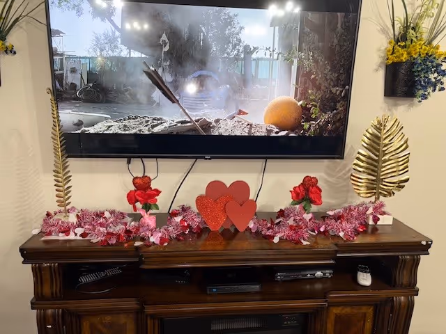 A wooden entertainment console with decorative Valentine's Day themed items including red and pink flowers, heart-shaped ornaments, and two gold leaf sculptures on either side. Above the console is a mounted flat-screen TV displaying a scene with an arrow stuck in a pumpkin outdoors.