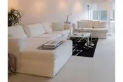 A bright and spacious living room featuring two large white sectional sofas arranged around a glass coffee table on a black area rug with a white geometric pattern. There is a potted plant in the corner and large windows allowing natural light to fill the room.