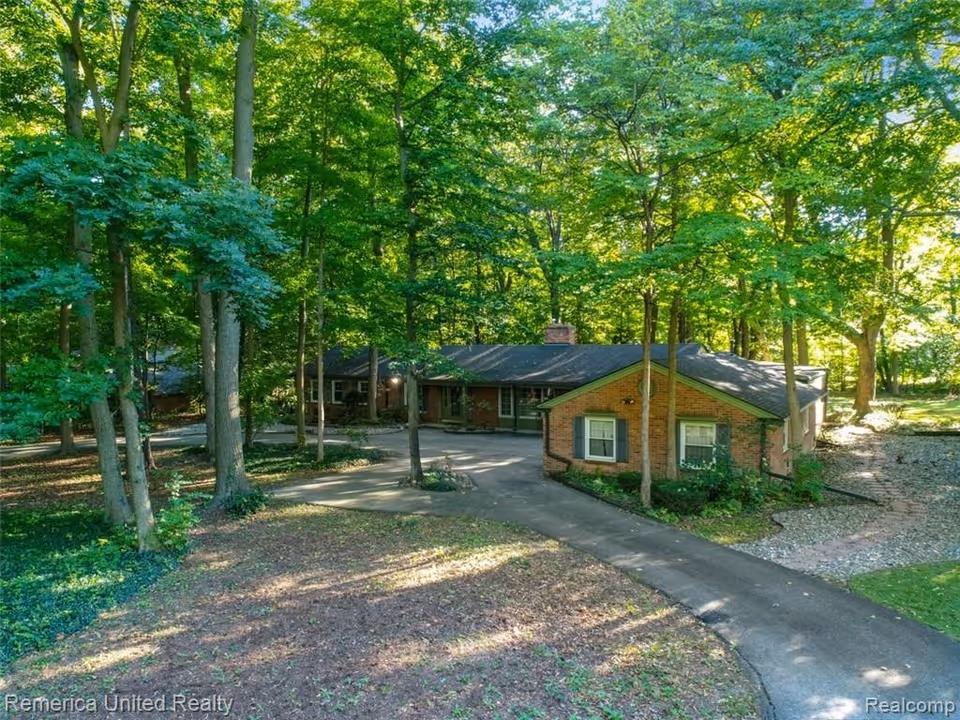A single-story brick house surrounded by tall green trees with a paved driveway leading to the entrance, situated in a wooded area with sunlight filtering through the leaves.