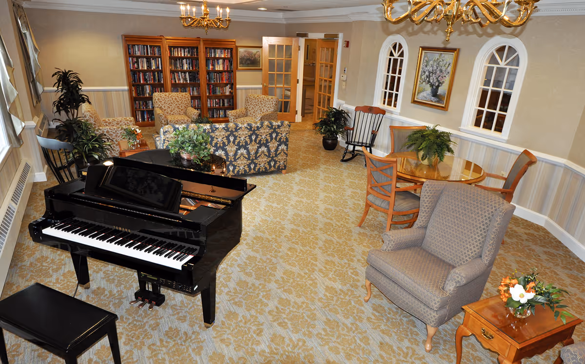 Spacious senior living common room with a grand piano, upholstered seating, bookshelves, and a small dining table.