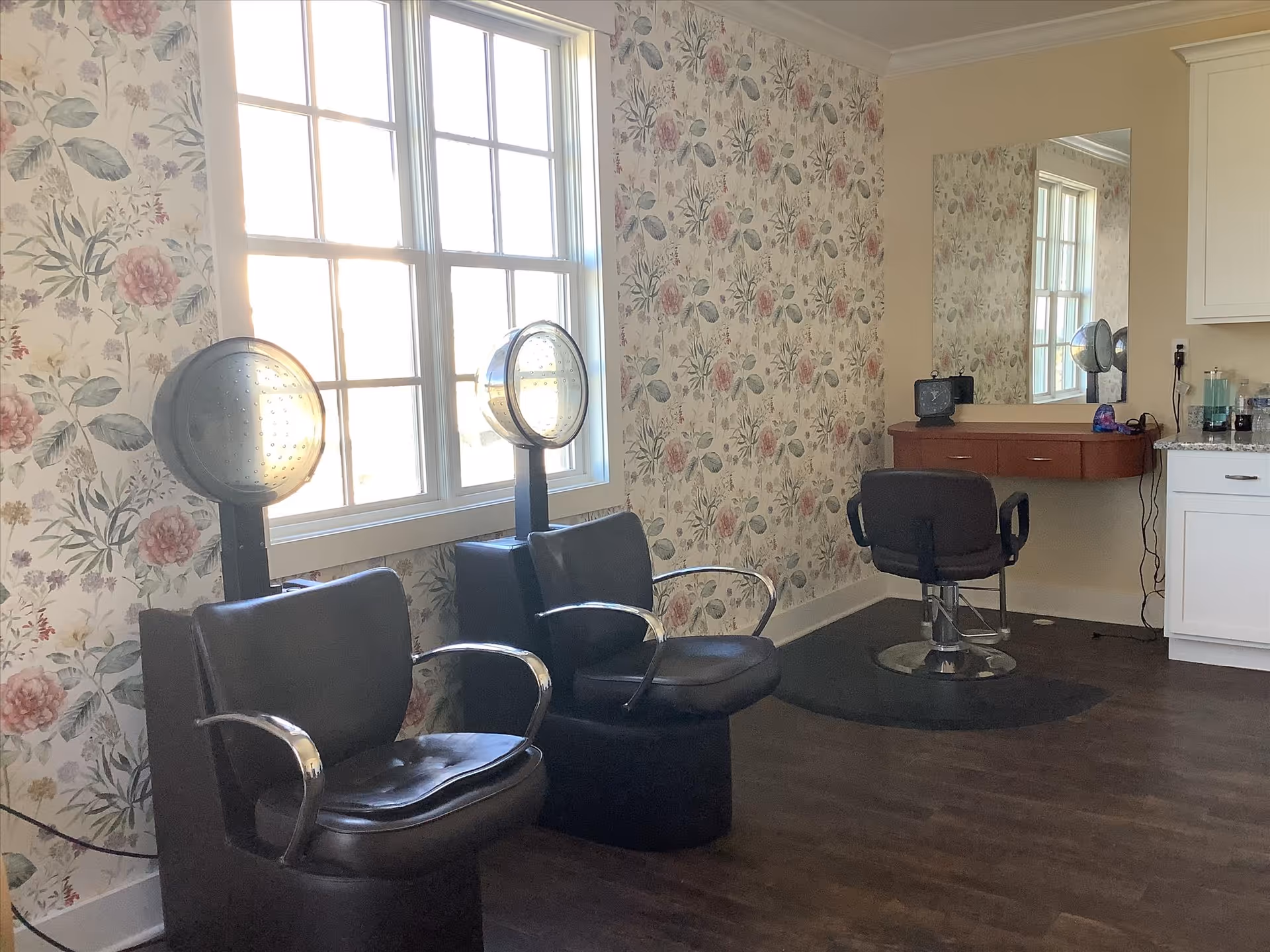 A salon-style room with two hooded hair dryers, three black styling chairs, a large wall mirror, floral wallpaper, and a window.