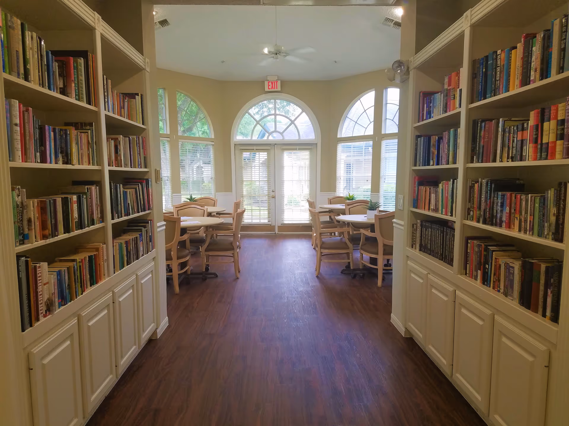 Interior view of a library or reading room in a senior living facility with bookshelves filled with books on both sides of a hallway leading to a seating area with round tables and chairs near large arched windows and a glass door.