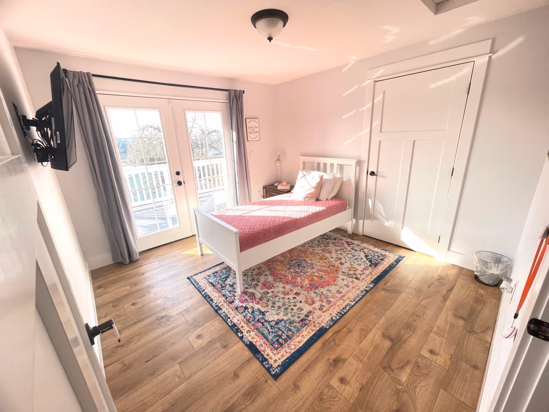A bright bedroom with a single white bed featuring a pink quilt and pillows, a colorful patterned rug underneath, wooden flooring, a wall-mounted TV, and double glass doors with gray curtains leading to an outdoor balcony.