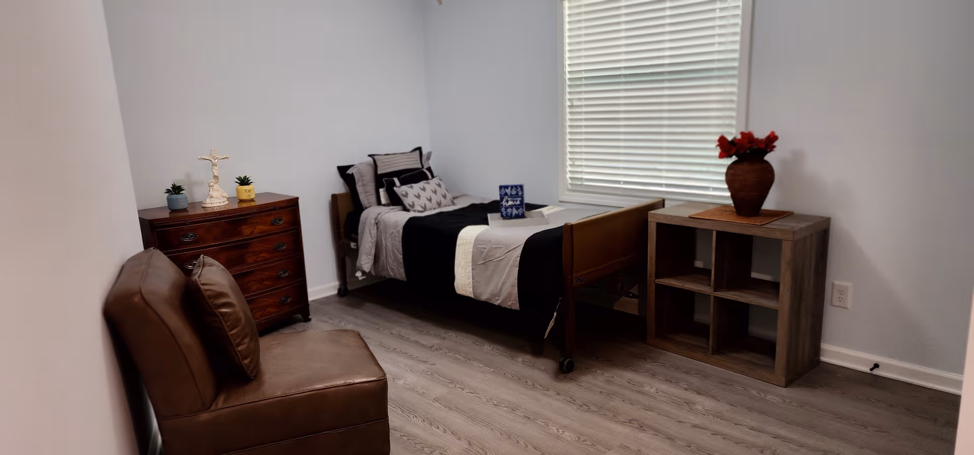 A simple senior care bedroom with a single bed made with gray and black bedding, a wooden dresser with small plants and a decorative item, a brown leather chair, and a wooden cube shelf with a vase of red flowers near a window with closed blinds.