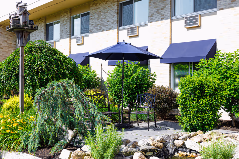 Courtyard patio with metal chairs and a navy umbrella surrounded by shrubs, a small rock pond, and the building facade with window awnings.