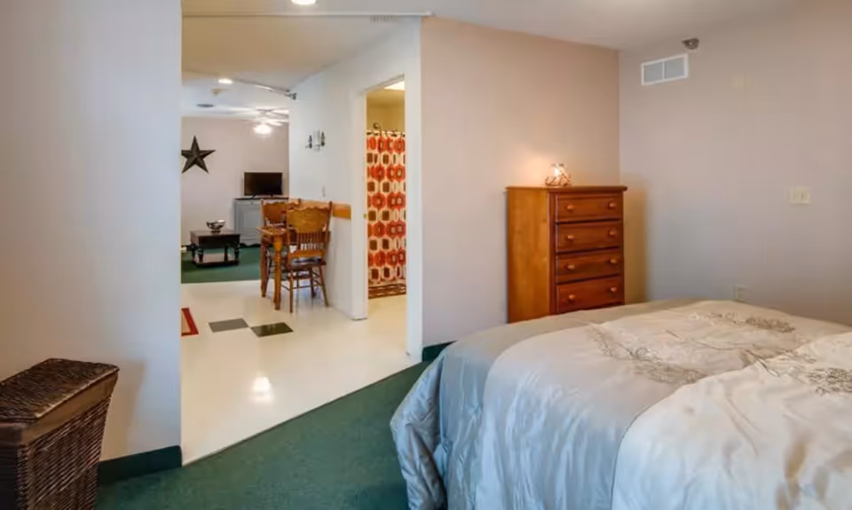 View of a senior living facility room showing a bed with a light-colored comforter, a wooden chest of drawers, and an open doorway leading to a dining area with a table and chairs, a television, and a bathroom with a shower curtain featuring a red and white pattern.