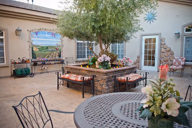 Outdoor patio area with a central raised planter containing a tree and flowers, surrounded by cushioned benches. Metal chairs and a round metal table with a flower arrangement are in the foreground. The background shows building walls with windows, a door, a mural of a vineyard landscape, and garden tools on a rack.