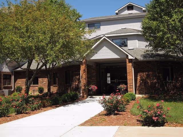 Front entrance of a brick senior living building with a covered portico, walkway, trees, and flowering shrubs.