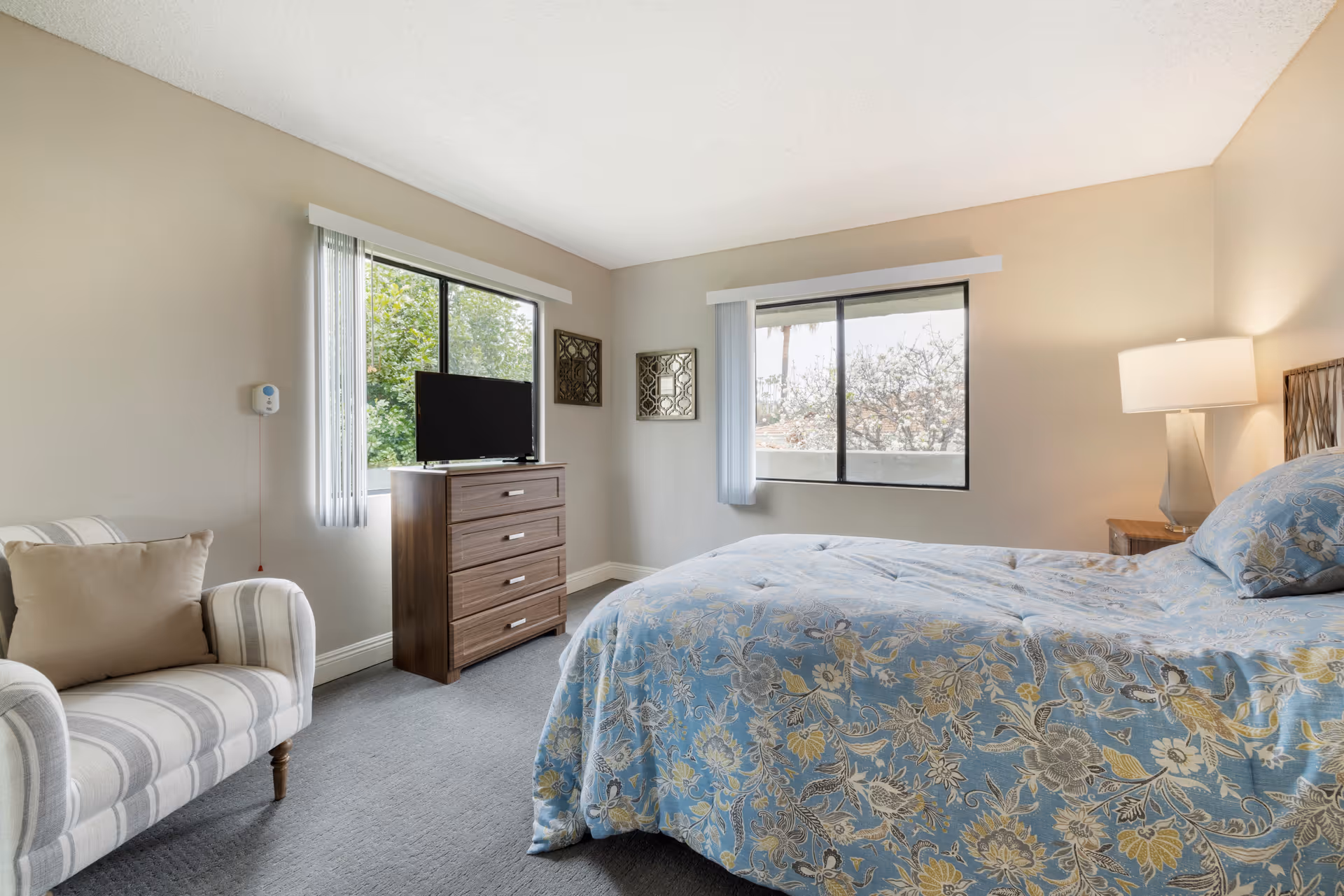 A bright and cozy bedroom in a senior living facility featuring a bed with a blue floral bedspread, a wooden nightstand with a lamp, a striped armchair with a beige pillow, a wooden dresser with a flat-screen TV, and two windows with vertical blinds letting in natural light.