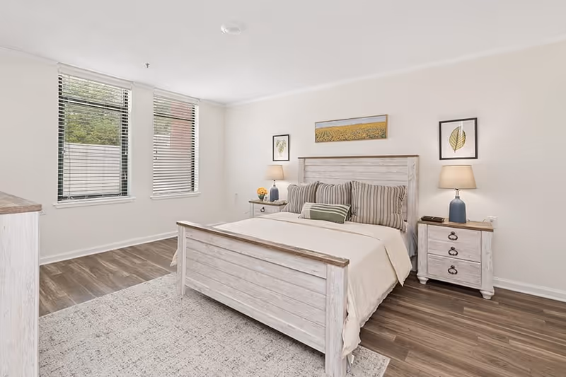 A bright bedroom with a wooden bed frame and matching nightstands on either side. The bed is made with beige bedding and several pillows, including striped and solid green ones. Two table lamps with blue bases and beige shades sit on the nightstands. Above the bed are three framed artworks featuring botanical themes. The room has two large windows with blinds, wood flooring, and a light-colored area rug partially under the bed.