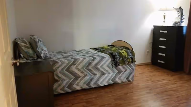 A simple bedroom with a single bed covered in a chevron-patterned bedspread and two pillows. There is a dark wooden nightstand next to the bed and a matching tall dresser with a lamp and decorative item on top. The room has wooden flooring and plain white walls.
