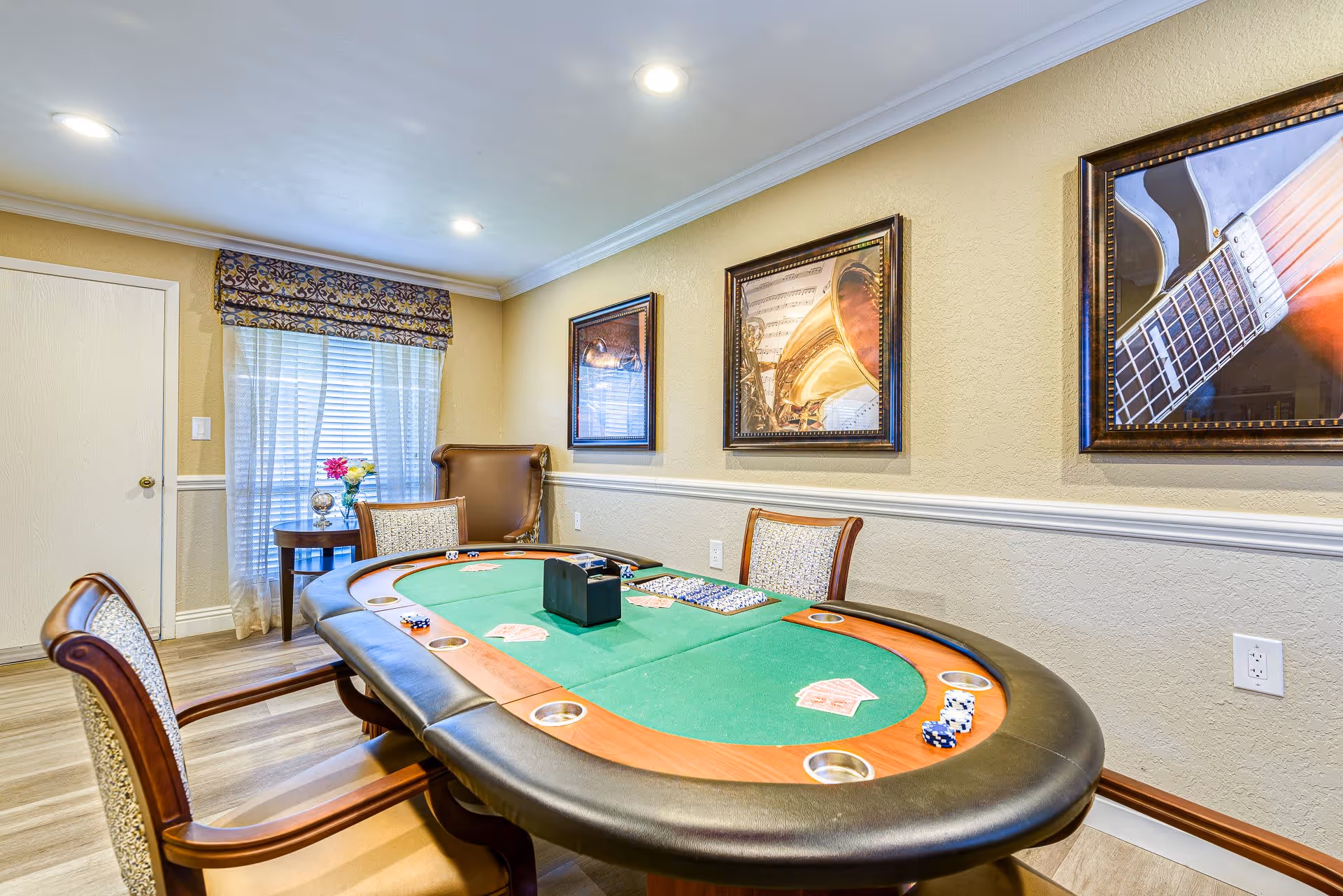 A well-lit room with a poker table set up with playing cards and poker chips. The room has beige walls with white trim, three framed pictures of musical instruments on the wall, a window with sheer curtains and a patterned valance, and several wooden chairs with cushioned seats around the table.