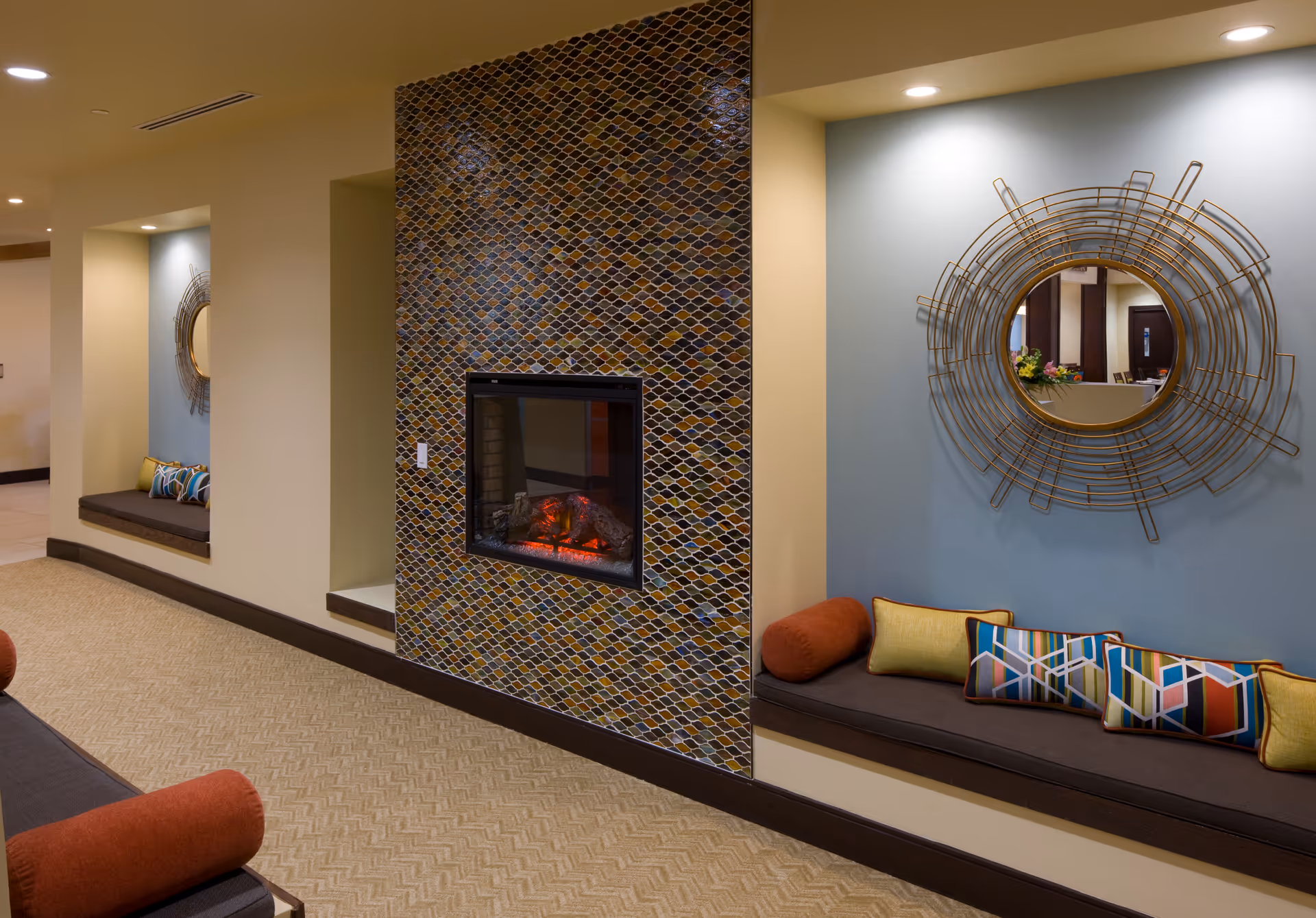 Cozy communal lounge featuring a tiled fireplace, built-in bench seating with colorful cushions and a decorative round mirror.