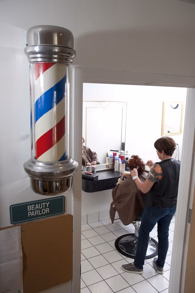View into a beauty parlor room where a hairstylist is cutting or styling a seated client's hair. A traditional barber pole is mounted on the wall outside the room next to a sign that reads 'Beauty Parlor'. The room has white tiled floors, a large mirror, and various hair care products on the counter.