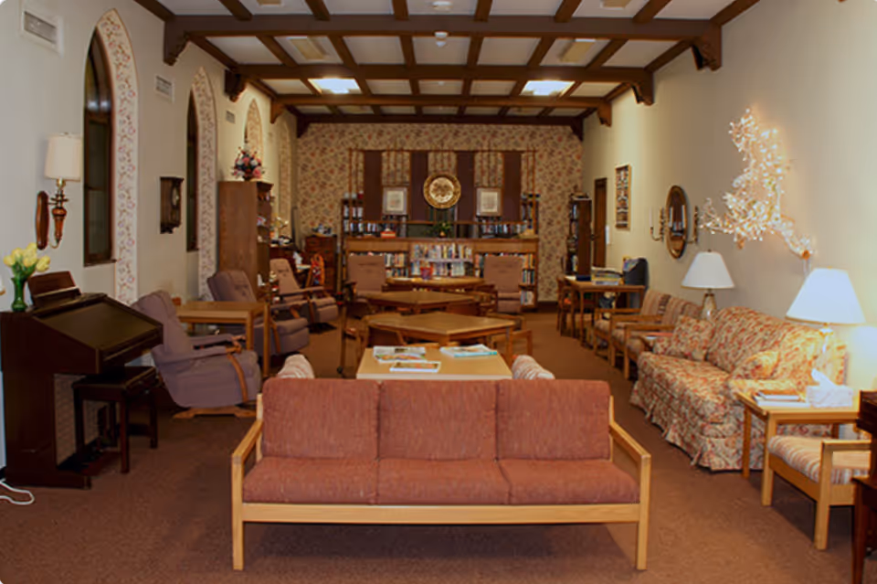 A cozy living room in a retirement home with multiple seating options including a red cushioned sofa, floral patterned couch, armchairs, and wooden tables. The room features a wooden coffered ceiling, floral wallpaper on the far wall, bookshelves filled with books, and decorative wall lamps. There is a piano on the left side and a unique light fixture on the right wall.