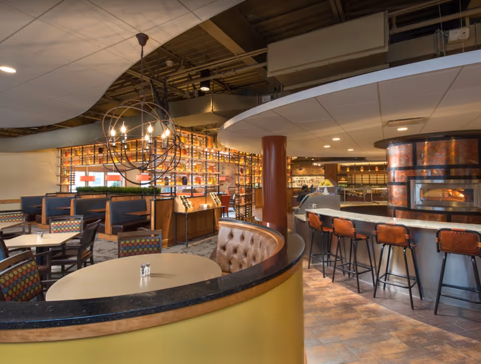 Interior view of a dining area in Garden Spot Village featuring round and rectangular tables with chairs, a curved booth with tufted leather seating, a bar with high stools, a modern chandelier, and a wood-fired oven. The space has a warm and inviting atmosphere with exposed ceiling ducts and decorative shelving.